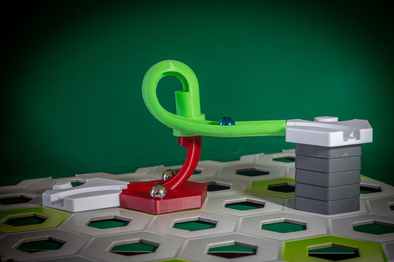 Puede incluir: Una pista de m&aacute;rmol verde y roja con una torre blanca y gris. La pista est&aacute; hecha de pl&aacute;stico y tiene un bucle. La torre est&aacute; hecha de bloques apilados. La pista est&aacute; colocada sobre un tablero hexagonal blanco y verde.