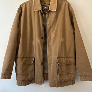 May include: A brown jacket with a corduroy collar and two large front pockets. The jacket has a button-up closure and a plaid lining. The jacket appears to be made of a durable, worn material.