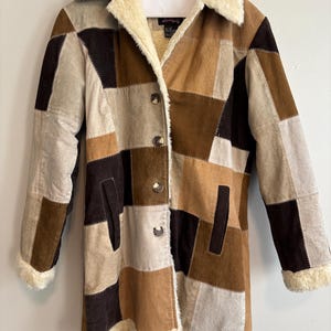 Puede incluir: Una chaqueta de gamuza con parches de color marrón, beige y negro con cuello y forro de piel sintética. La chaqueta tiene cierre de botones y dos bolsillos.