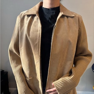 May include: A tan suede jacket with a collared neckline and a full-length zipper. The sleeves are a ribbed knit material. The jacket has two front pockets and a tailored fit. The jacket is a neutral color and can be worn in many settings.