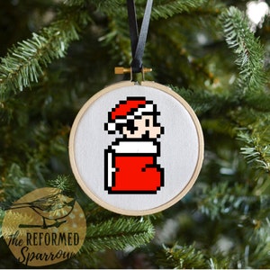 May include: A white embroidery hoop with a red and white pixelated image of a video game character wearing a Santa hat and holding a stocking. The character is standing in front of a white background.