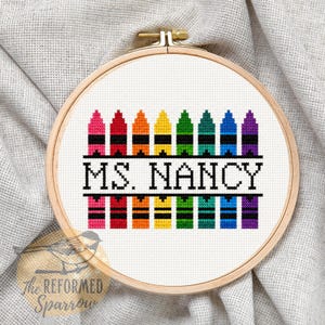 May include: A circular wooden embroidery hoop containing white fabric with a cross-stitch design. The design features two rows of colorful crayon illustrations in pink, red, orange, yellow, green, teal, blue, and purple, with the text MS. NANCY in black.