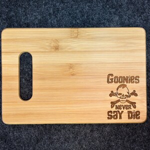 Peut inclure: Une planche &agrave; d&eacute;couper en bois avec une poign&eacute;e. La planche est grav&eacute;e au laser avec un motif de cr&acirc;ne et d'os crois&eacute;s avec le texte "Goonies Never Say Die".