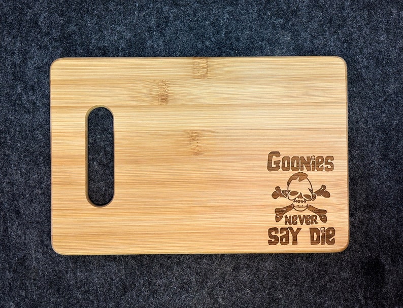 Peut inclure: Une planche &agrave; d&eacute;couper en bois avec une poign&eacute;e. La planche est grav&eacute;e au laser avec un motif de cr&acirc;ne et d'os crois&eacute;s avec le texte "Goonies Never Say Die".