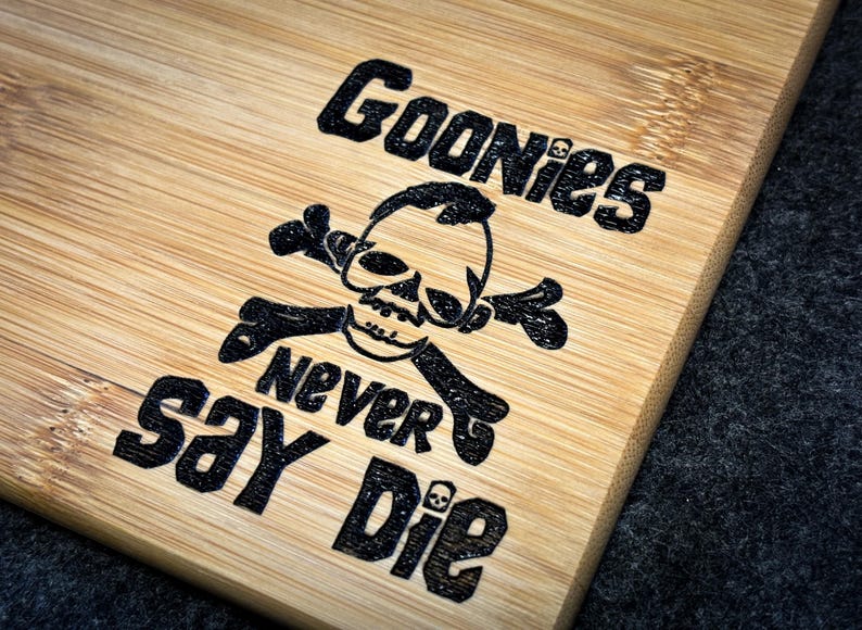 Peut inclure: Une planche &agrave; d&eacute;couper en bois avec un motif noir et blanc repr&eacute;sentant un cr&acirc;ne et des os crois&eacute;s, les mots "Goonies Never Say Die" et un motif de grain de bambou.