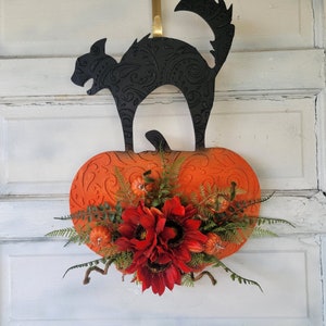 Könnte beinhalten: Eine schwarze Katzensilhouette auf einem orangefarbenen Kürbis mit einem Blumenarrangement aus roten Sonnenblumen und grünen Blättern. Der Kürbis und die Katze sind an einem goldenen Aufhänger befestigt.