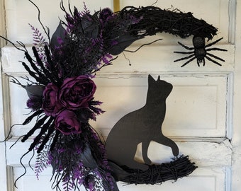 Puede incluir: Una corona de luna negra en forma de media luna con flores moradas, ramitas negras y una silueta de gato negro. Una araña negra está posada en la corona.