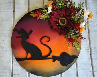 Puede incluir: Decoración redonda de Halloween de madera con silueta de gato negro en escoba sobre fondo degradado de atardecer. Adornada con girasoles burdeos, flores blancas, amarillas y rojas, y vegetación. Una pieza festiva para decoración.