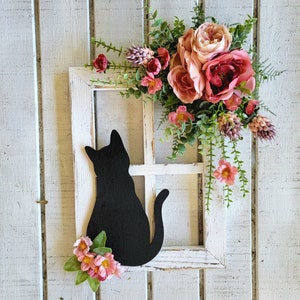 May include: A decorative wooden cat silhouette sits within a white window frame, adorned with pink and peach-colored flowers and greenery. The black cat is perched, with a curved tail. The frame is distressed, giving it a rustic appearance.