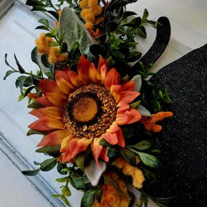 Puede incluir: Un colgador de puerta negro con purpurina con un girasol, vegetaci&oacute;n y tallos amarillos y esponjosos.