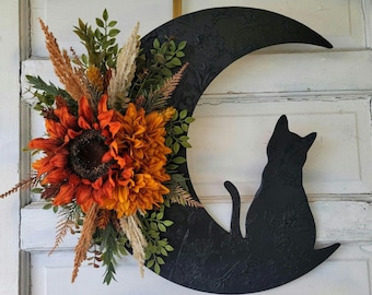 Puede incluir: Una luna de madera negra en forma de media luna con una silueta de gato negro sentada sobre ella. La luna está decorada con una corona floral de otoño con girasoles naranja, amarillo y marrón.