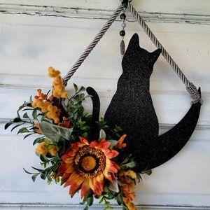 Puede incluir: Una silueta de gato negro con detalles de purpurina est&aacute; colgada en una puerta. El gato est&aacute; sentado en una luna creciente y est&aacute; decorado con un girasol y vegetaci&oacute;n.