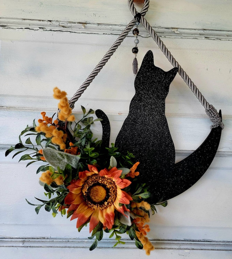 Puede incluir: Una silueta de gato negro con detalles de purpurina est&aacute; colgada en una puerta. El gato est&aacute; sentado en una luna creciente y est&aacute; decorado con un girasol y vegetaci&oacute;n.