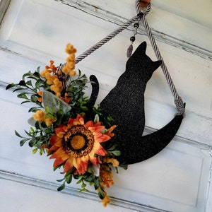 Puede incluir: Una silueta de gato negro en una luna creciente con una corona de girasol y vegetaci&oacute;n colgando de una puerta blanca.