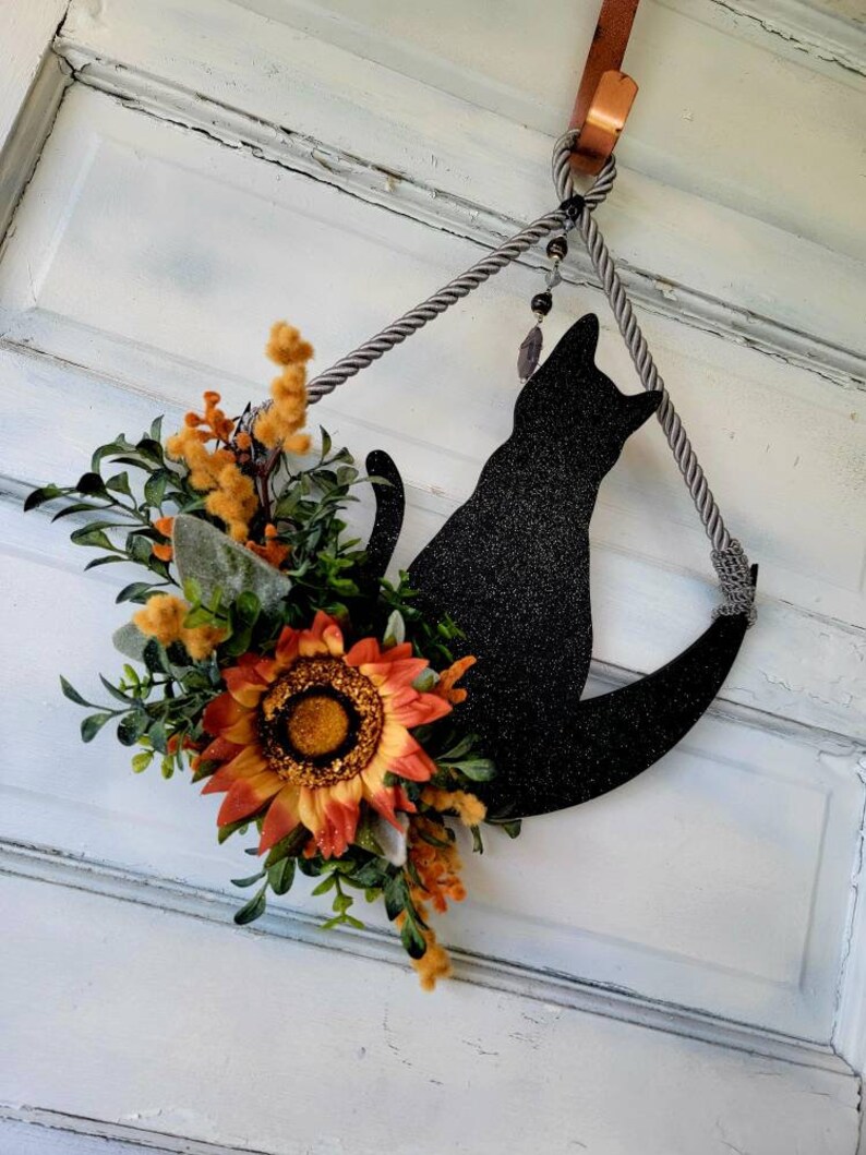 Puede incluir: Una silueta de gato negro en una luna creciente con una corona de girasol y vegetaci&oacute;n colgando de una puerta blanca.