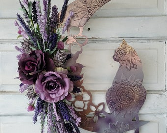 Puede incluir: Una luna creciente de metal de color púrpura y dorado con una silueta de gato y un diseño floral. La luna está decorada con flores de lavanda y moradas.