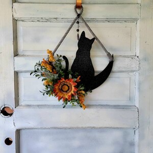 Puede incluir: Una silueta de gato negro con una luna creciente, decorada con un girasol y follaje de oto&ntilde;o, cuelga de una puerta blanca con un gancho de cobre.