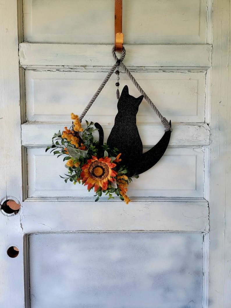 Puede incluir: Una silueta de gato negro con una luna creciente, decorada con un girasol y follaje de oto&ntilde;o, cuelga de una puerta blanca con un gancho de cobre.