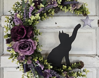Puede incluir: Una corona en forma de media luna con una silueta de gato negro que alcanza una estrella gris. La corona está adornada con rosas moradas, lavanda y follaje verde. Cuelga de una puerta de madera blanca desgastada.