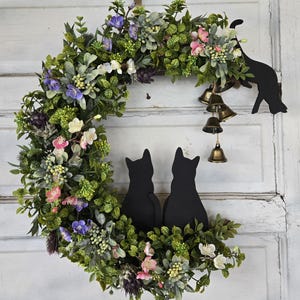 Op de afbeelding: Een halvemaanvormige krans met weelderig groen, paarse, roze en witte bloemen. Twee zwarte katten silhouetten zitten aan de basis, met een katten silhouet en messing bellen die van bovenaf hangen. Een charmant, decoratief stuk.
