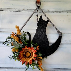 Puede incluir: Una silueta de gato negro con purpurina, colgada de una cuerda gris con un abalorio de cuentas. El gato est&aacute; sentado en una forma de media luna. Un arreglo floral de oto&ntilde;o con girasoles y vegetaci&oacute;n est&aacute; unido a la parte inferior de la luna.