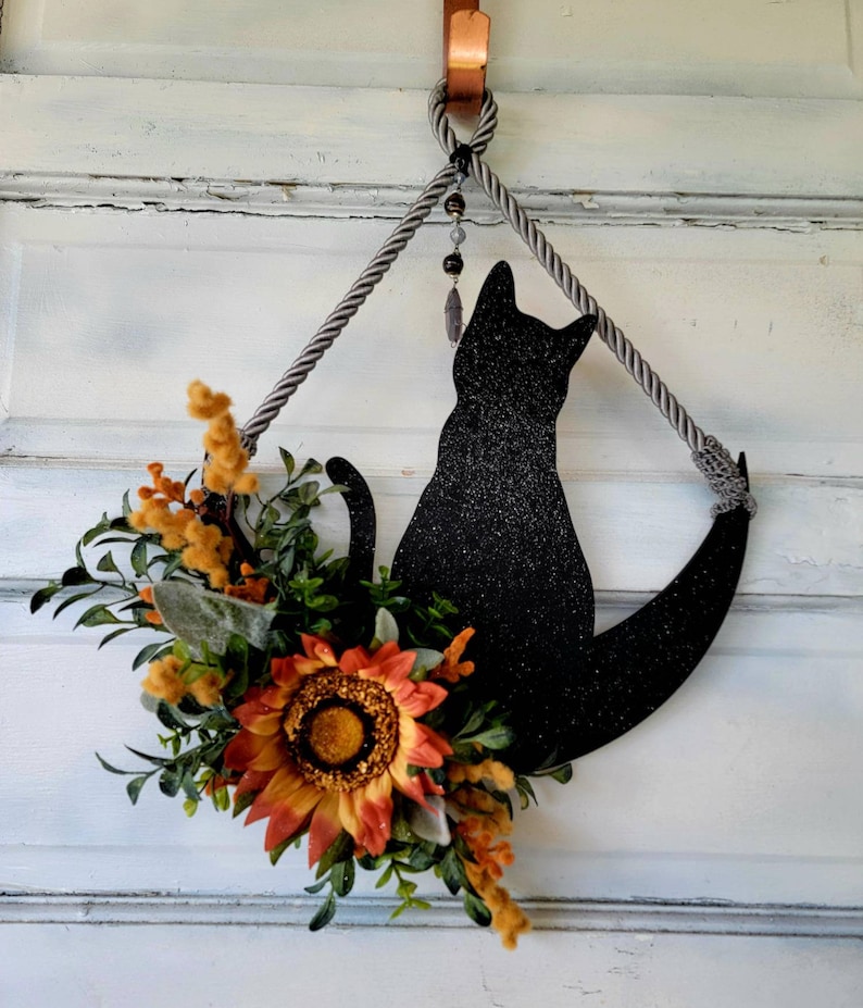 Puede incluir: Una silueta de gato negro con purpurina, colgada de una cuerda gris con un abalorio de cuentas. El gato est&aacute; sentado en una forma de media luna. Un arreglo floral de oto&ntilde;o con girasoles y vegetaci&oacute;n est&aacute; unido a la parte inferior de la luna.