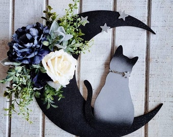May include: Black crescent moon with a white cat silhouette sitting on it. The moon is decorated with silver glitter and small silver stars. The cat has a silver glitter collar with a star charm. The moon is surrounded by a floral arrangement of white roses and blue hydrangeas.