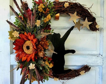 May include: A fall-themed wreath featuring a black cat silhouette reaching for gold stars. The wreath is made of grapevine and decorated with sunflowers, autumn leaves, and other floral accents.