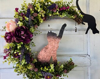 Puede incluir: Una silueta de gato de metal negro con un amuleto colgante cuelga de una corona de flores moradas, rosas y verdes. La corona está hecha de flores y vegetación artificiales.
