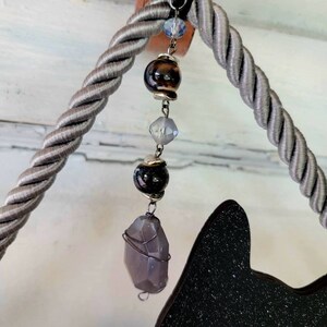 Puede incluir: Una silueta de gato negro con purpurina, colgada de una cuerda gris con una cadena de cuentas de cristal y piedras preciosas.