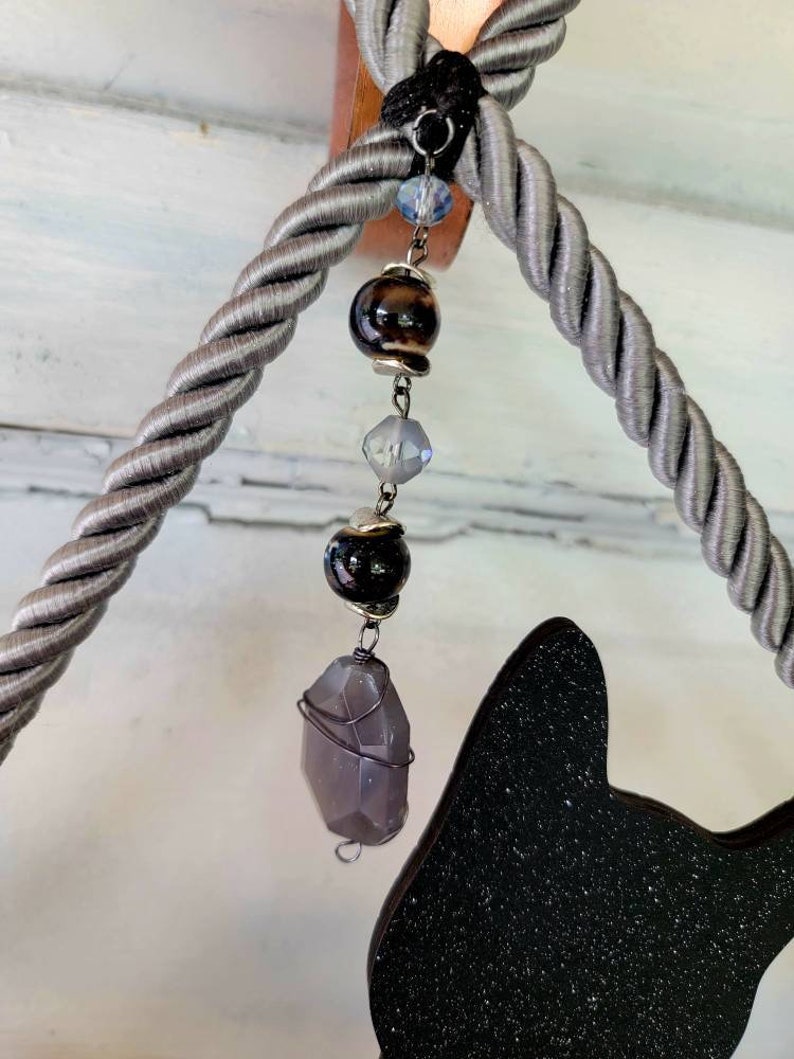 Puede incluir: Una silueta de gato negro con purpurina, colgada de una cuerda gris con una cadena de cuentas de cristal y piedras preciosas.
