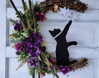 May include: A purple and green floral wreath with a black cat silhouette reaching for white stars. The wreath is made of grapevine and has a brown ribbon hanging from the top.