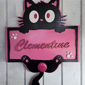 May include: A pink wooden sign with a black cat peeking over the top. The sign says "Clementine" in pink cursive lettering. The cat has white eyes and red pupils. The sign has white paw prints on either side of the name.