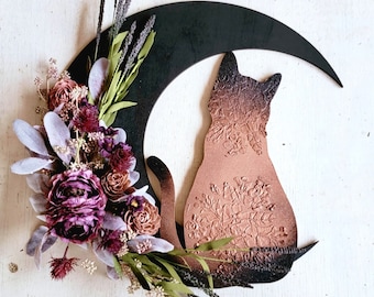 Puede incluir: Una luna creciente negra con una silueta de gato de color cobre, decorada con un arreglo floral de flores moradas y marrones, hojas verdes y ramitas de lavanda.