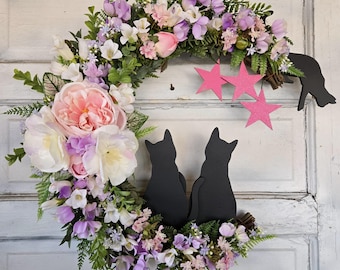 May include: A floral wreath shaped like a crescent moon with two black cat silhouettes and pink stars. The wreath is made of artificial flowers in shades of pink, white, and purple.