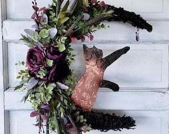 Puede incluir: Una corona de ramas negras con forma de media luna con una silueta de gato de cobre y flores moradas. La corona está decorada con vegetación y un colgante de cristal morado.
