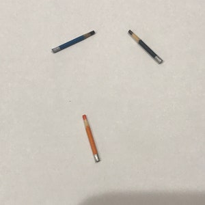 May include: Three miniature pencils in blue, black, and orange, each with a silver ferrule. The pencils are arranged on a white surface, with the blue pencil at the top left, the black pencil at the top right, and the orange pencil at the bottom.