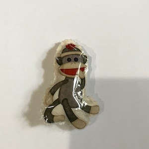 May include: A gray and brown fabric monkey with a red hat and a white background. The monkey is wearing a red scarf and has a white face with black eyes.