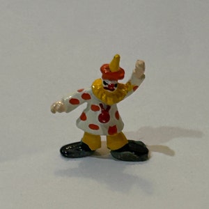May include: A small, white clown figurine with red polka dots, a yellow ruffle collar, and a red hat with a yellow point. The clown is painted with a red nose and black eyes and is waving with its right arm.
