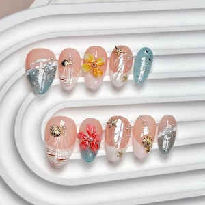 Op de afbeelding: Een set van tien kunstnagels met een strandthema. De nagels zijn geschilderd in verschillende kleuren, waaronder roze, wit, blauw en goud. Sommige nagels hebben 3D bloemen- en schelpenontwerpen, terwijl andere glitter- en golfpatronen hebben.