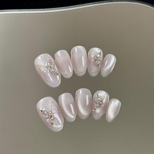 Peut inclure: Un ensemble de dix faux ongles avec une finition œil-de-chat scintillante dans un ton gris lavande pâle. Les ongles de forme ovale sont ornés de breloques papillon irisées et de grappes de petits strass transparents et étincelants.