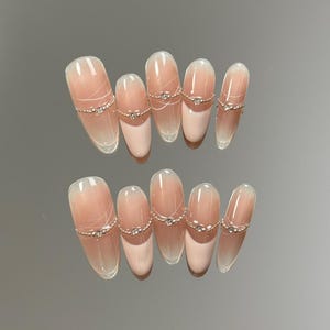 Op de afbeelding: Een set van tien ovaalvormige kunstnagels. Elke nagel heeft een gradiëntontwerp, dat overgaat van lichtroze naar een heldere punt. Delicate zilverkleurige kettingen met kleine, heldere strass-steentjes zijn op de nagels aangebracht en voegen een vleugje elegantie toe.