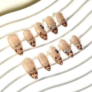 May include: A set of ten almond-shaped false nails. The nails are a nude colour with a leopard print design on the tips. Some nails have gold accents and rhinestones.