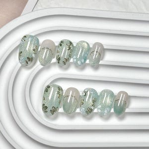 May include: A set of oval false nails. The nails feature light blue and glittery green bases with floral patterns. Some nails are solid light pink with a gold stripe. The nails are arranged in two rows.