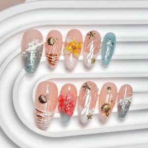May include: A set of ten almond-shaped press-on nails featuring a beach theme. The designs include nude bases with 3D elements like gold seashells, starfish, white pearls, and sculpted flowers in yellow and pink, alongside blue glitter tips.