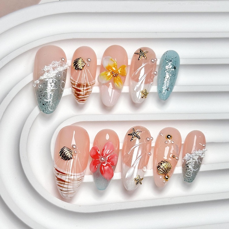May include: A set of ten almond-shaped press-on nails featuring a beach theme. The designs include nude bases with 3D elements like gold seashells, starfish, white pearls, and sculpted flowers in yellow and pink, alongside blue glitter tips.