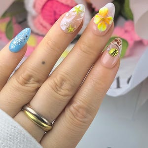 May include: Almond-shaped nails featuring a variety of designs including a light blue base with silver glitter, a nude base with white swirls, gold starfish and pearl accents, a yellow floral 3D element, and a gold seashell charm on a nude base.