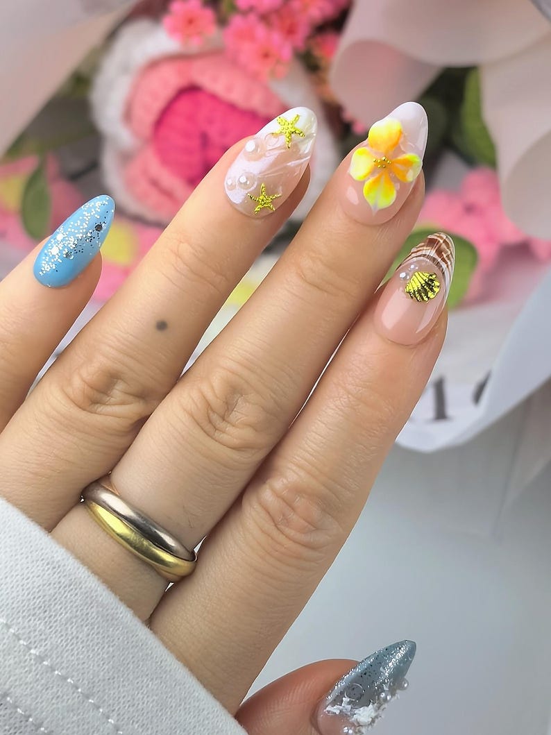 May include: Almond-shaped nails featuring a variety of designs including a light blue base with silver glitter, a nude base with white swirls, gold starfish and pearl accents, a yellow floral 3D element, and a gold seashell charm on a nude base.