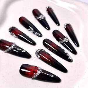 Puede incluir: Diez uñas postizas brillantes. Cada uña presenta un diseño degradado, pasando de un rojo oscuro en la base a negro en la punta. Algunas uñas están adornadas con brillantes pedrería transparente, añadiendo un toque de glamour.
