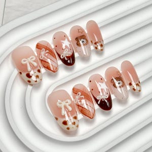 May include: A set of ten almond-shaped press-on nails featuring a nude base with brown accents. Designs include raised white bows, brown plaid patterns, brown polka dots, small brown bear faces, and white star shapes with a glossy finish.